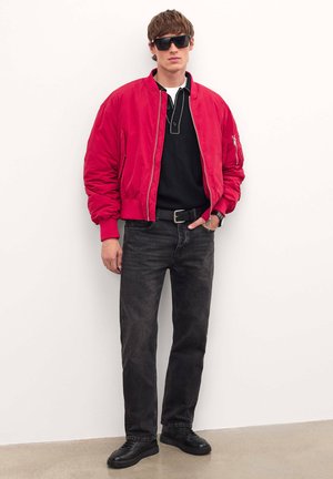 Rote Bomberjacke, schwarzes Polohemd, schwarze Jeans und schwarze Sneakers. Accessoires umfassen Sonnenbrillen und eine Uhr. Glattes Material, lässige Passform.