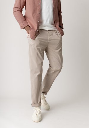 Mann trägt beige Chinos mit hochgekrempelten Säumen, weiße Sneaker, weißes T-Shirt und ungeknöpftes rosa Cordhemd, steht mit Händen in den Taschen.