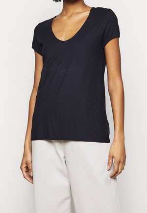 Vrouw draagt een zwart T-shirt met korte mouwen en V-hals en lichte broek tegen een effen achtergrond.