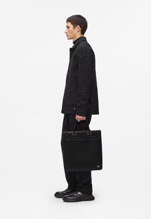 Black tote bag with rectangular shape, smooth leather texture, dual handles, and subtle logo detail. Model wearing a textured black outfit.