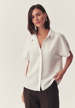 Kobieta o ciemnych włosach, nosząca złote kolczyki, luźną białą koszulę zapinaną na guziki z krótkim rękawem oraz ciemne spodnie, stojąca na jednolitym tle.