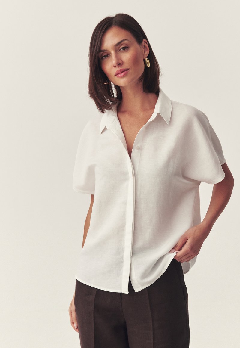 Femme aux cheveux foncés portant des boucles d'oreilles dorées, une chemise blanche à manches courtes et boutons, ample, et un pantalon foncé, debout devant un fond uni.