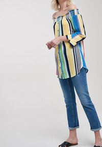 Stripet off-the-shoulder skjorte i gul, blå og pink, med knapper ned langs foran og blondedetaljer ved halsen. Bæres med opbrettede jeans.