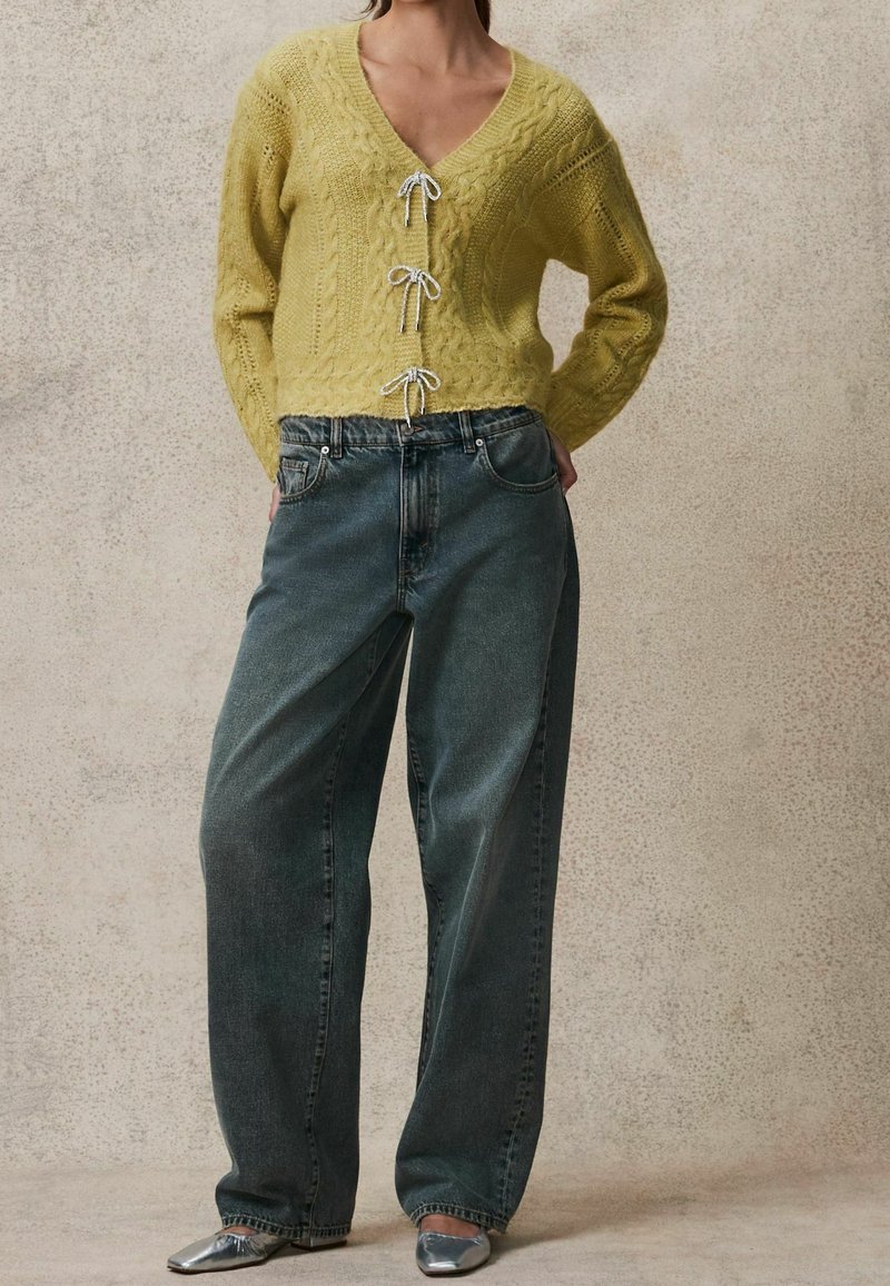 Personne portant un cardigan court jaune en maille torsadée avec trois nœuds noués, un jean large gris foncé, et des chaussures argentées à bouts pointus.