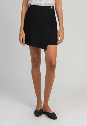 Femme portant une jupe portefeuille asymétrique noire avec une épingle en forme d'étoile et des ballerines pointues noires, debout devant un fond uni.