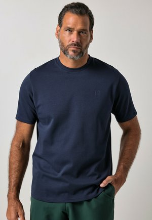 Uomo adulto con barba e capelli corti indossa una camicia blu scuro tinta unita a maniche corte e pantaloni verdi, in piedi con una mano in tasca.