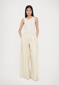 Vaalean beige leveälahkeiset housut, joissa on laskokset ja sivutaskut, yhdistettynä istuvaan off-white väriseen ribbimäiseen tanktoppiin, jossa on nappikoristus.