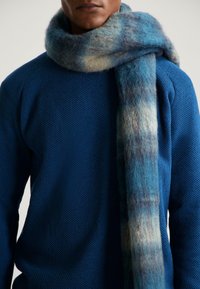 Man in een gestructureerde blauwe sweater met een dikke, zachte, blauw en grijs geblokte sjaal om de nek gewikkeld.