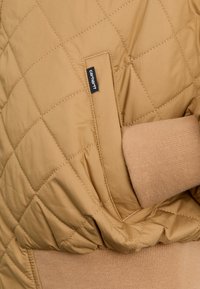 Chaqueta acolchada de color marrón claro con una cintura acanalada y detalles en los bolsillos. Presenta una etiqueta de marca negra en el lado superior izquierdo.
