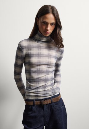 Joven mujer con cabello largo y oscuro que lleva un jersey de cuello alto de cuadros gris y beige y vaqueros azul oscuro con un cinturón marrón, de pie frente a un fondo liso.