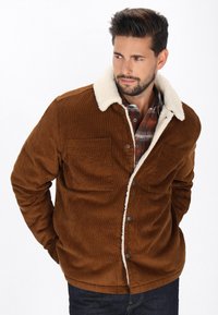 Braune Cordjacke mit weißem Sherpa-Kragen, ausgestattet mit zwei Fronttaschen und Knopfverschluss, getragen über einem karierten Hemd.