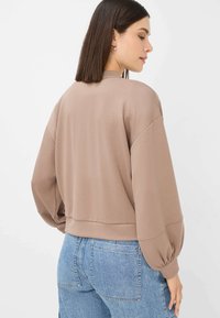 Brown pullover sweatshirt with dropped shoulders and balloon sleeves. Soft texture with a cropped fit. Paired with light blue denim.