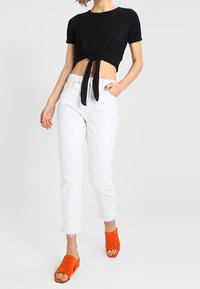 Černé krácené tričko s mašlí, bílé džíny s vysokým pasem a oranžové semišové sandály s otevřenou špičkou a chunky podrážkou.