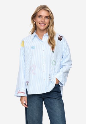 Smilende kvinde med langt blondt hår iført en løs blå- og hvidstribet skjorte med farverige broderede mønstre og blå jeans.