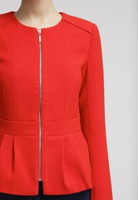 Veste rouge à manches longues avec fermeture éclair argentée à l'avant, taille cintrée et ourlet plissé portée par une personne sur un fond clair uni.