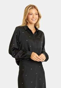 Czarna satynowa bluzka z kołnierzykiem, ozdobiona rozrzuconymi srebrnymi ćwiekami, z długimi, bufiastymi rękawami z mankietami oraz luźnym krojem.