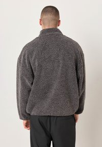 Uomo di spalle che indossa una giacca in pile grigio scuro testurizzata e pantaloni neri su uno sfondo chiaro uniforme.