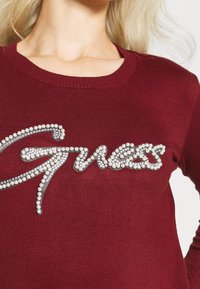 Burgundská svetr s žebrovaným kulatým výstřihem a ozdobným logo 'Guess' vyzdobeným průhlednými korálky a flitry.