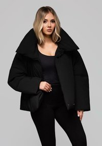 Černá péřová bunda s přeplněným límcem, s matným povrchem a zipem, nošená přes černý top a legíny.