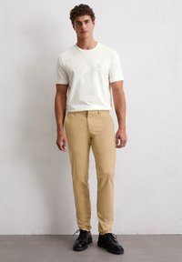 Biała bawełniana koszulka z logo na piersi, zestawiona z beżowymi spodniami slim-fit i czarnymi sznurowanymi butami. Gładka tekstura i codzienny styl.