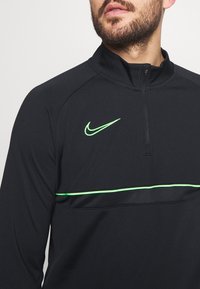 Musta neljännesvetoketjullinen urheilupaita korkealla kauluksella. Edessä vihreä Nike-logo ja vihreä korostevyö rinnan kohdalla. Sileä materiaali.