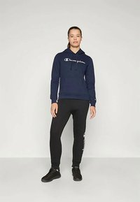 Námořnická modrá mikina s kapucí a bílým logem, v kombinaci s černými těsnými tepláky. Oba vyrobené z měkkého materiálu; tepláky mají logo detail po straně.