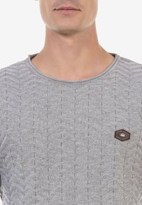 Grauer gestrickter Pullover mit einem strukturierten Zickzackmuster, Rundhalsausschnitt und einem kleinen Emblem auf der Brust. Weicher Stoff und figurbetonter Schnitt.