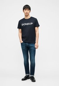 Άνδρας που φοράει μαύρο t-shirt με το λογότυπο "DONDUP", μπλε τζιν κατεβασμένα μέχρι τον αστράγαλο και μαύρα παπούτσια, στέκεται με το ένα χέρι στην τσέπη μπροστά σε απλό φόντο.