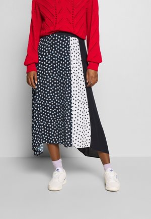 Woman wearing a color-block midi skirt with black, white, and blue polka dots, a red sweater, lilac socks, and white sneakers.