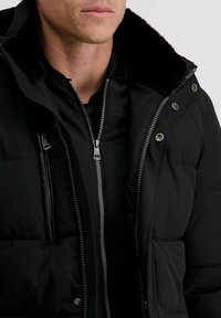 Man in een zwart gewatteerd jack met rits, een bontkraag en zichtbare metalen ritsen en drukknoopjes aan de voorkant.