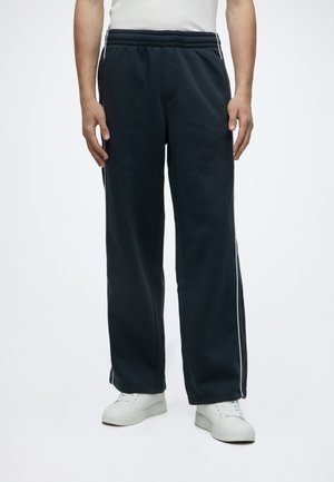 Mand iført sorte løstsiddende sweatpants med hvide sidelommer og hvide sneakers, stående mod en ensfarvet lys baggrund.