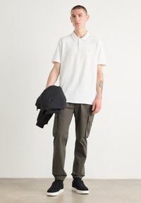 Hvit polo skjorte med krage, olivenfarge cargo bukser og svarte sneakers. Modellen holder en svart jakke. Polert stoff og avslappet passform.