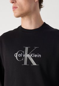 Jaunas vyras dėvi juodą megztinį su dideliu pilku "CK" ir baltu "Calvin Klein" logotipu ant krūtinės, žiūrintis į kairę.