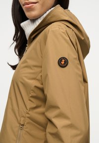 Γυναίκα που φοράει μπεζ μπουφάν με κουκούλα, με μαύρο και πορτοκαλί κυκλικό λογότυπο "Save the Duck" στο πάνω μέρος του μπράτσου, πάνω από λευκό ριμπ πλεκτό ζιβάγκο.