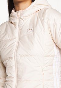 Světlá krémová péřová bunda se zipem a kapucí. Obsahuje bílé boční pruhy a malé vyšívané logo na hrudi.