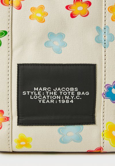 Lerrets totebag med flerfargede blomstmønstre, beige bakgrunn, og en svart etikett som viser merke, stil, sted og årstall.