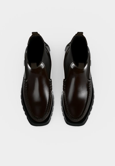 Paire de bottes en cuir lisse marron foncé, sans lacets, avec des semelles épaisses noires, vues de dessus sur un fond blanc uni.