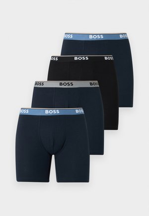 Fiatal, csinos testalkatú férfi áll mezítláb, fekete, BOSS márkájú boxernadrágot visel, egyszerű világos háttér előtt.