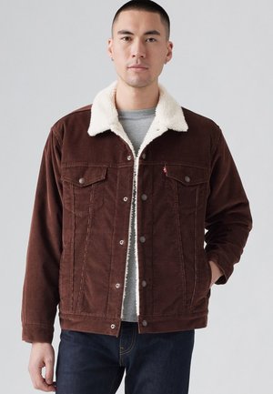 Chaqueta de pana marrón con un cuello de piel sintética color crema, bolsillos frontales y botones a presión; llevada sobre una camisa gris, combinada con jeans oscuros.