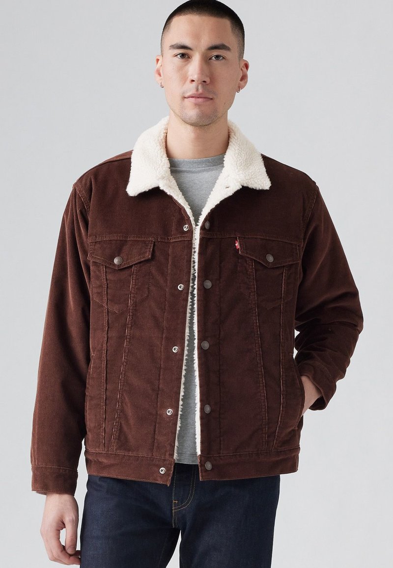 Bruine corduroyjas met een crèmekleurige nepbontkraag, voorzakken en drukknoopsluiting; gedragen over een grijze shirt, gecombineerd met donkere denim.