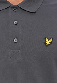 Pilkas polo marškinėliai, pasižymintys tekstūruota medžiaga, trimis juodais mygtukais ir siuvinėtu geltonu erelio logotipu kairėje krūtinės pusėje.