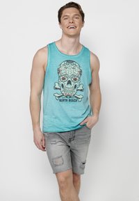 En turkos linne har en detaljerad skallegrafik med blommiga accenter och korsben, i kombination med slitna grå shorts.