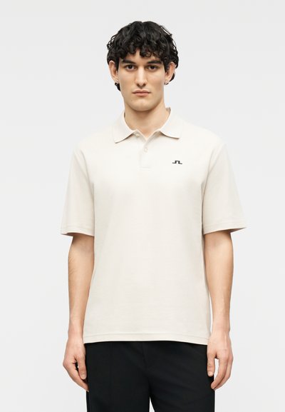 Jeune homme aux cheveux courts et bouclés portant un polo beige clair avec un petit logo sombre sur le côté gauche de la poitrine et un pantalon noir.