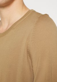 Pulover tricotat de culoare maro deschis, cu guler rotund, margini ribbate și o textură netedă, evidențiind designul fără cusături la umeri.