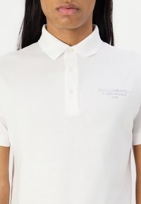 Bijela polo majica od pamučnog materijala s klasičnim ovratnikom i tri dugmeta. Sadrži utisnuti logotip "Karl Lagerfeld" na prsima.
