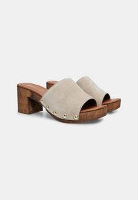 Mules en daim à bout ouvert de couleur beige clair avec un talon en bois. Dotées de clous dorés le long de la sangle. Semelle plate pour le confort.