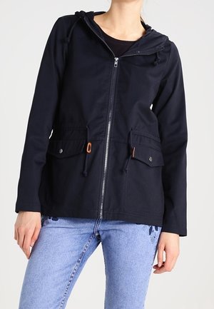 Woman wearing a black zip-up hooded jacket with front flap pockets and light blue jeans with floral embroidery.