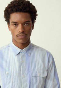 Šviesiai mėlyna ir balta dryžuota marškiniai su klasikiniais atlapais ir dviem krūtinės kišenėmis; glotni, audinėmedvilnės medžiaga.