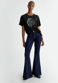 Maglietta nera con stampa circolare bianca. Jeans flared in denim scuro con cuciture a contrasto. Modello in piedi contro uno sfondo chiaro.