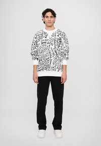White sweatshirt with black abstract text pattern, ribbed cuffs and hem. Paired with black pants and white sneakers. Model standing upright.